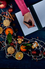 A girl with a red manicure in a knitted pink sweater writes a list of cases for the new year. A list of Christmas presents. Typical festive winter accessories, attributes, ornaments.
