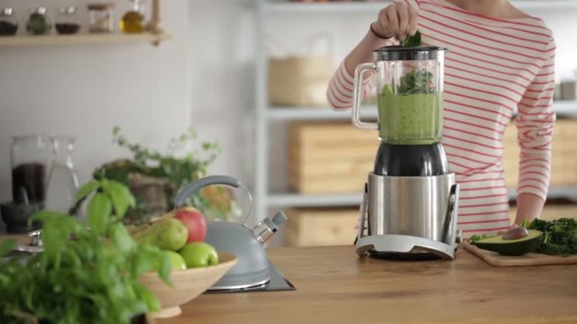 Close-up of a woman making a healthy green smoothie with spinach and avocado in a blender on wooden countertop. Slide video.
