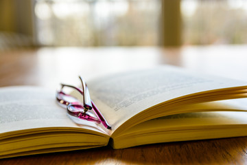 Book and glasses on wooden table. Close-up view of an open pocket book with a pair of reading glasses on it, laying flat on a wooden table in front of a window.