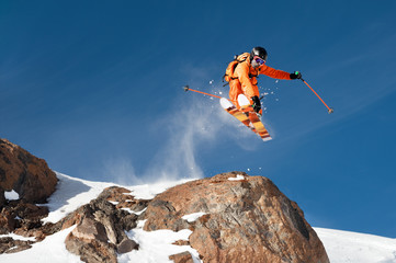 A professional skier makes a jump-drop from a high cliff against a blue sky leaving a trail of snow...