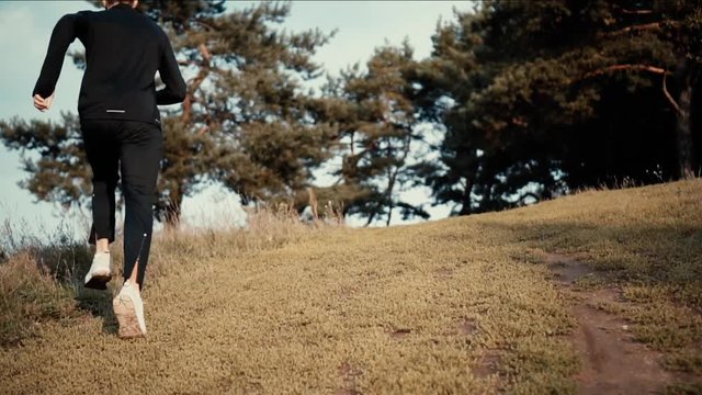 Slow motion runner going uphill to a forest. Low angle back view. Young athlete exploring fall countryside landscapes.