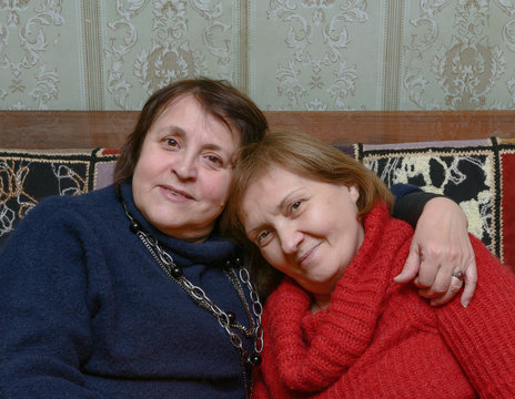 Two aged women are sitting embracing on the sofa.