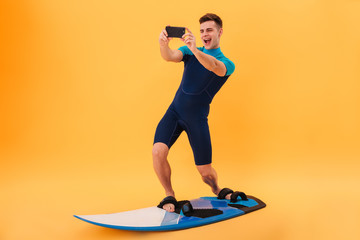 Full length photo of young smiling man in swimsuit taking selfie on smartphone while surfing on surfboard