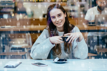 Happy woman drinking coffee in cafe in cold winter day. Model, holding cup of coffee, looking at you and smailing. View through the window. Snowfall. Christmas, new year, winter holidays concept.