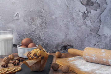 Chocolate muffin, a glass of milk, organic eggs in a white bowl, rolling pin, flour sprinkled on a wooden board, dried oranges, walnuts, anise and a stone plate on a brown wooden table