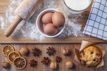Top view on chocolate muffin, organic milk in a glass, eggs in a white bowl, rolling pin on a wooden board sprinkled with flour, dried oranges, anise, walnuts and a linen napkin in a checkered pattern