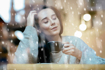Cute dreamy woman sitting in cozy cafe, drinking hot coffee in snowy winter day. Model closing eyes. Photo taken through the window. Magic, fairy tale concept. Snowfall effect.