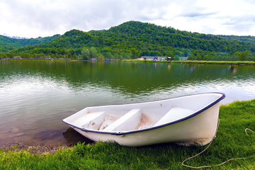 A boat without oars tied with a rope is on the green grass in front of the pond on the background of the countryside and the forest mountain landscape. White boat on the lake shore, Sochi, Russia.