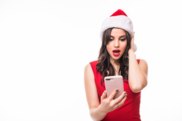 Portrait of a shocked pretty caucasian girl dressed in red christmas hat holding mobile phone while standing and looking at camera over white background