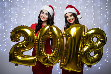 Fototapeta na wymiar Portrait of two women in santa hat and red dress with 2018 numbers in hands og New Year on bokeh light background.