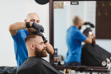 Fototapeta na wymiar Male professional hairdresser serving client by scissors. Ginger handsome brutal stylish young man with thick big beard, short hair getting trendy haircut in black cape. Light white barber shop room.