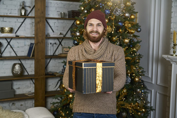 Fototapeta premium A young man gives gift to his partner for the new year or Christmas male model in trendy sweater and hat hipster against the backdrop of christmas tree