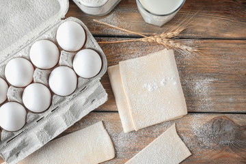 Raw flaky dough and carton of eggs on wooden table, top view
