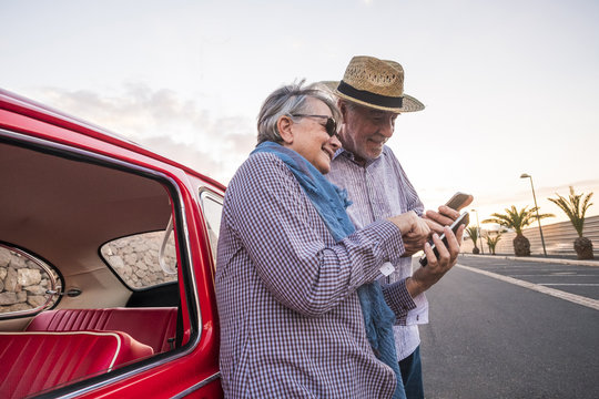 Elderly Couple With Hat, With Glasses, With Gray And White Hair, With Casual Shirt, On Vintage Red Car On Vacation Enjoying Time And Life. With A Cheerful Mobile Phone Smiling