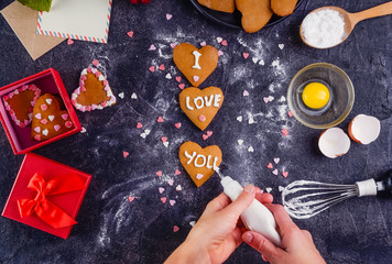 Top view female hands decorating homemade cookies in shape of heart as gift for lover on Valentine's day. Dark stone background with ingredients, gift box and decor. Selective focus. Copy space