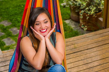 Outdoor view of smiling young beautiful woman relaxing in a hammock with both hands under her chin, in wooden nature background
