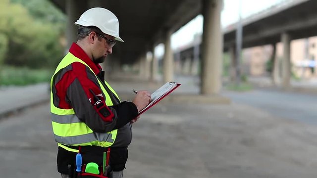 Road mender makes notes in work documentation. Building inspector with work documentation at project site under overpass