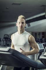 Fototapeta na wymiar Blond attractive young man running on treadmill, looking at camera