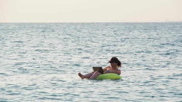 Girl with a laptop on an inflatable circle in the sea. Happy girl with a laptop floating on the waves.