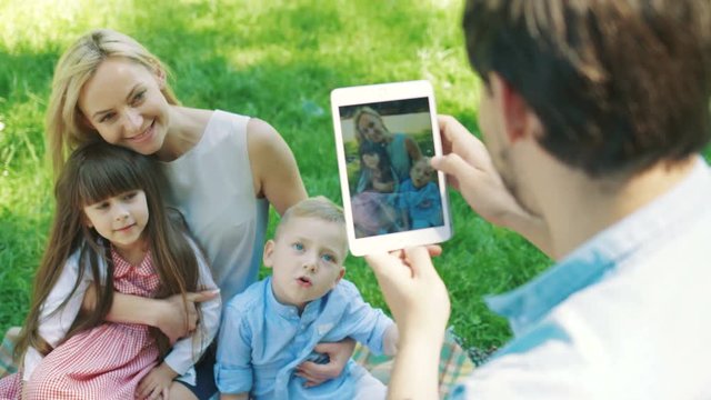 Rear of caucasian father taking a photo on the tablet computer of his blonde beautiful wife with two kids sitting on the grass in the park in summer. Sunny weather. Outdoor