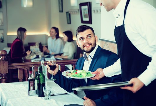 Waiter serving delicious salads to handsome young man at restaurant