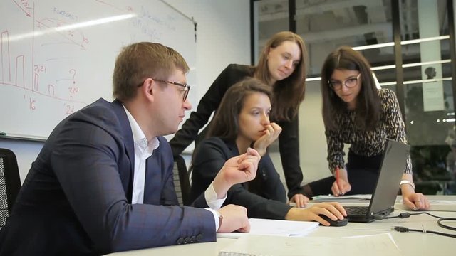 Head and staff brainstorm in front of laptop in conference room. An adult man in glasses and young women look at screen and make decision consulting each other.