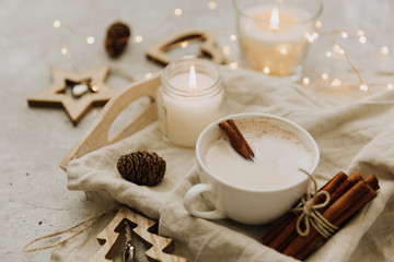 Cup of milky cinnamon coffee on a tray with illuminated fairy lights, candles and festive Christmas ornaments