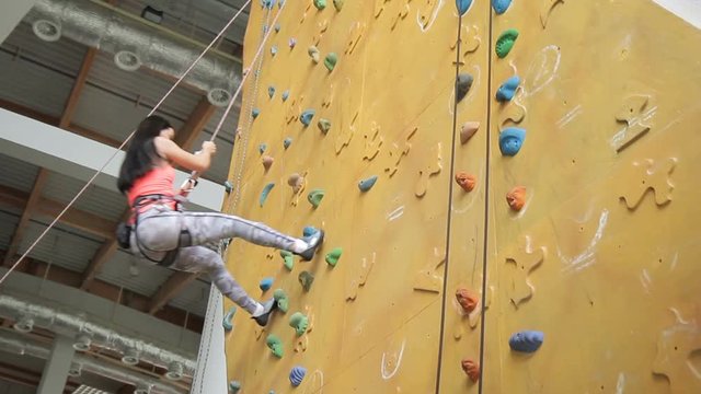 Young brunette goes down climbing wall in sports club. Woman in protective gear on body, holds to strong rope with hands and quickly descends, pushing with legs