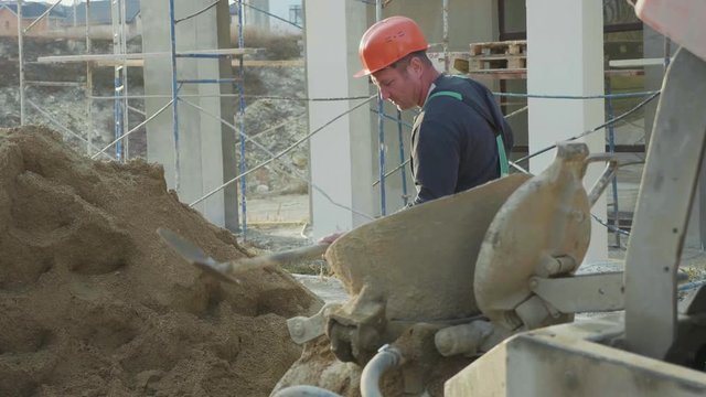 Caucasian worker pours sand into a concrete mixer by shovel at construction site