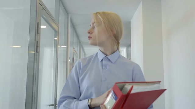 The portrait of blonde female secretary who is looking for her boss in the office. The manager in blue shirt is holdin the red folder with documents in her hands and turning her head while going