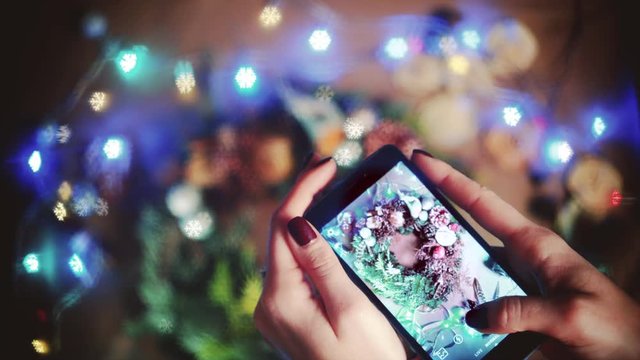 closeup female hands with phone making photo of handmade Cristmas wreath, colorful blurred background with figure bokeh