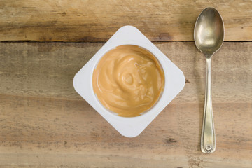 Healthy natural caramel creamy yoghurt in small white plastic cup on rustic wooden table with little spoon