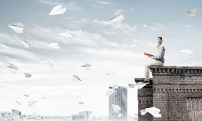 Handsome student guy on roof edge reading book and paper planes fly around