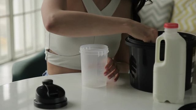 Young woman is putting protein into shaker while sitting home. Female is at table, opens plastic container and puts mixture from jar with spoon. Attractive brunette dressed in stylish top with bare