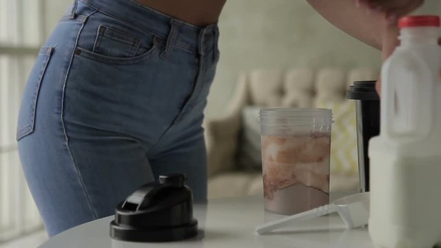 Young woman pours milk into shaker and shakes it at home. Female stands by table and puts fresh milk into plastic container with protein powder then closes lid and jiggles it carefully. Fitness lady