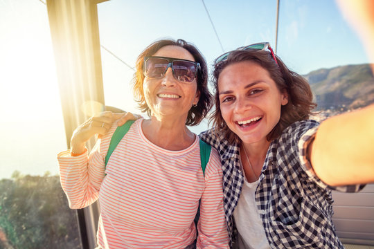 An elderly beautiful woman and her adult daughter travel together, do selfie on a mobile phone in the cockpit of a funicular during a summer holiday at sea - Powered by Adobe