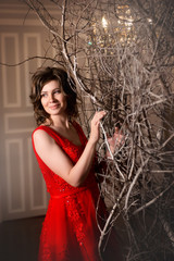 Brunette girl in red evening dress standing next to a decorative tree in vintage interiors