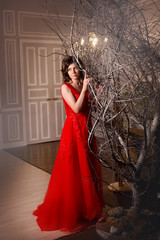 Brunette girl in red evening dress standing next to a decorative tree in vintage interiors