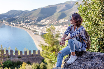 Naklejka premium young woman traveler with a backpack sitting and admiring the scenery with the sea and mountains at sunset