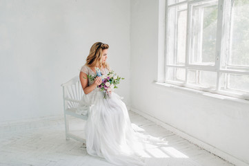 A beautiful bride in a white dress is sitting on a chair near the window and holding a wedding bouquet. White studio.