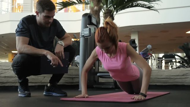 Male couch is explaining young female client how to do push ups in gym. The woman in pink t-shirt is standing on her knees and arma on the mat and the trainer in black sportswear is sitting near and