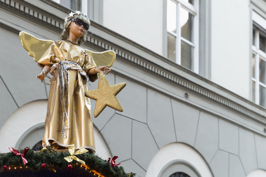 A statue of a cute little angel in a gold and silver robe, holding a star and wearing cool sunglasses on top of a christmas market booth.