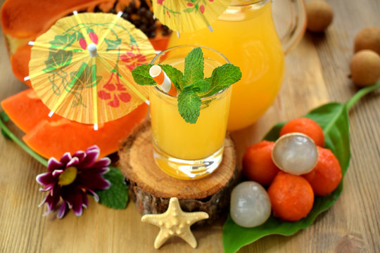 Yellow beverage decorated with mint, straws and little cocktail umbrellas in glass vessels surrounded by tropical fruits on a wooden table