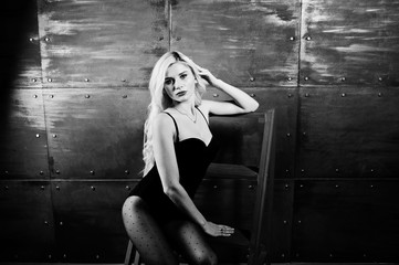 Elegante blonde girl wear on combi dress posed on chair against steel wall on studio.