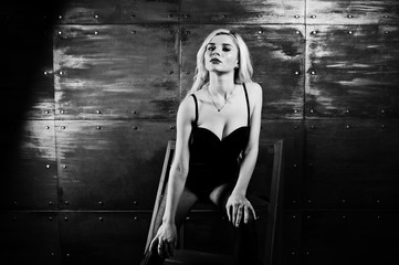 Elegante blonde girl wear on combi dress posed on chair against steel wall on studio.