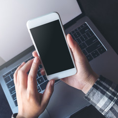 Mockup image of business woman holding mobile phone with blank black screen while using laptop on table in office