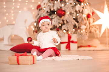 Obraz na płótnie Canvas Cute baby girl 1 year old wearing santa hat sitting over Christmas tree in room. Looking away. Winter season.