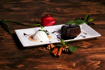 Close up of sweet chocolate brownie with vanilla ice cream, decorated with nuts, cinnamon and mint, served om white plate. Red tulip on wooden table