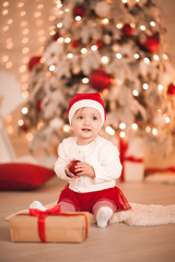Obraz na płótnie Canvas Smiling baby girl 1 year old sitting on floor with Christmas decorations over lights at background. Holiday season. Childhood.