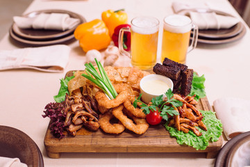 Plate of beer snacks, chips, fried chicken fillet, rusks, golden crispy squid rings, smoked pig ears, lettuce, green onions on wooden cutting board. Too beers on table